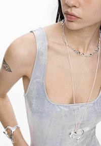 Hõbedast metallist kaelakee südamekujuga ripatsiga, kantud helehallil, keha järgival varrukateta topil, millel on sile ja veidi läikiv pind.