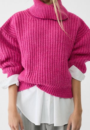 Pull à col roulé en grosse maille rose vif porté par-dessus une chemise blanche à boutons avec manches retroussées et pantalon gris.