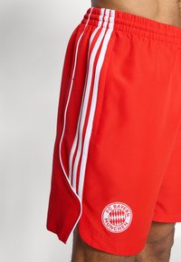 Piros sportos rövidnadrág fehér oldalsávokkal és rugalmas derékkal. Az alsó bal lábszáron a FC Bayern München logója látható.