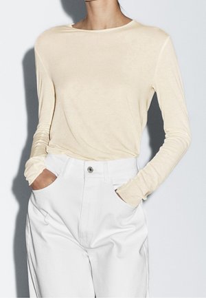 Persona che indossa una camicia beige a maniche lunghe infilata in pantaloni bianchi a vita alta con le mani nelle tasche, su uno sfondo semplice.