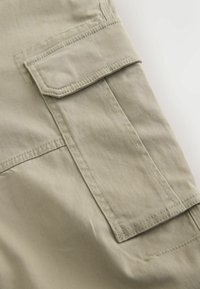 Nærbillede af beige stof cargo-lomme med klap og synlige syninger på tøj.