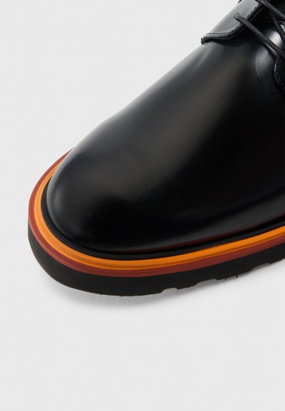 Zwarte leren schoen met een afgeronde neus, voorzien van een meerkleurige zool in lagen van oranje, rood en donkerbruin. Subtiele stikseldetails.