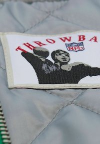 Grauer, gesteppter Stoff mit einem weißen Patch, der ein schwarz-weißes Grafikdesign einer Person zeigt, die einen Ball wirft, sowie rotem Text "THROWBA".