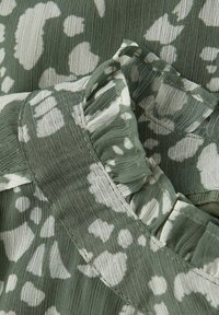 Zielony materiał z białym wzorem kwiatowym. Posiada falbaniaste brzegi i miękką, teksturowaną powierzchnię. Widoczne przeszycia wzdłuż szwów.