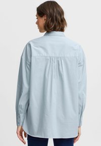 Light blue cotton button-up shirt with a back pleat, long sleeves, and buttoned cuffs, featuring a relaxed fit and smooth texture.