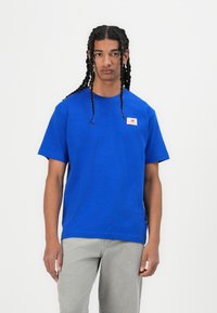 Blå bomulds t-shirt med rund hals og korte ærmer, med et lille logo-patch på venstre side. Model iført grå jeans.