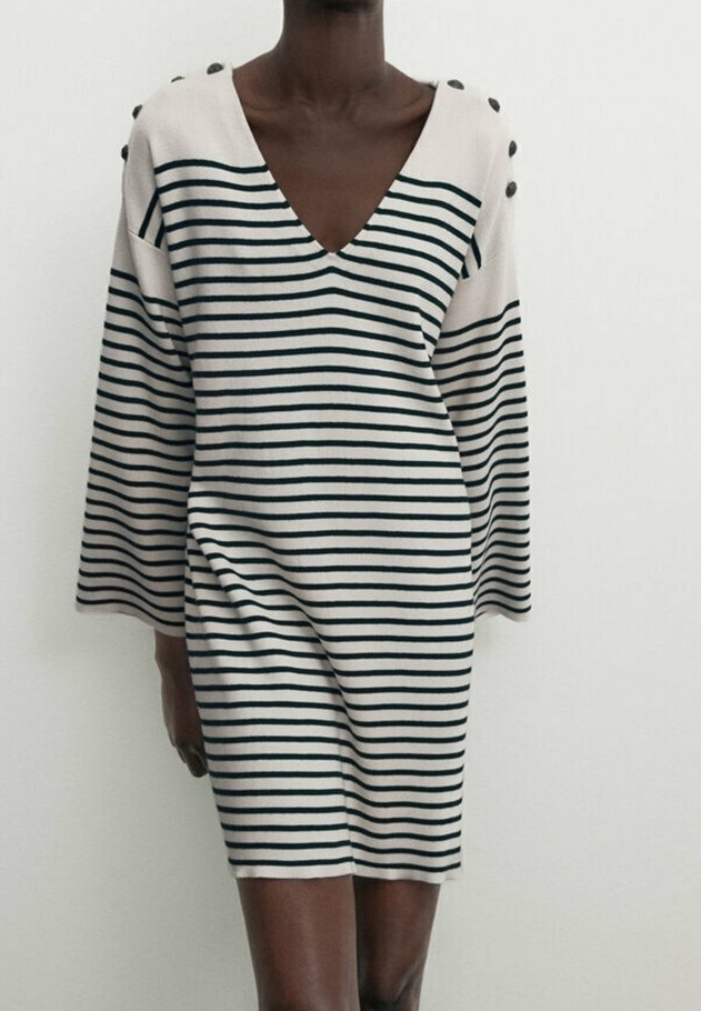 Personne portant une robe blanche à manches longues, longueur genou, avec de fines rayures horizontales noires et des boutons sur l'épaule.