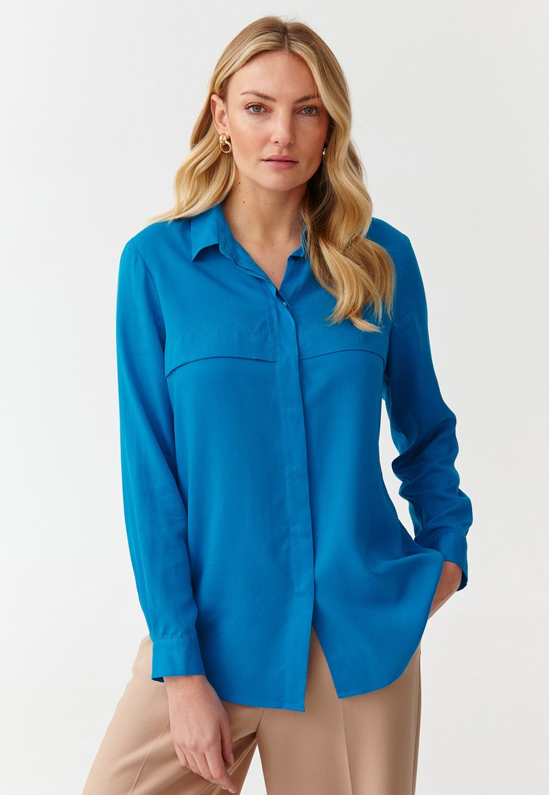 Blue blouse made of smooth fabric with a collar, long sleeves, and two front pockets. Plain design with a relaxed fit and curved hem.