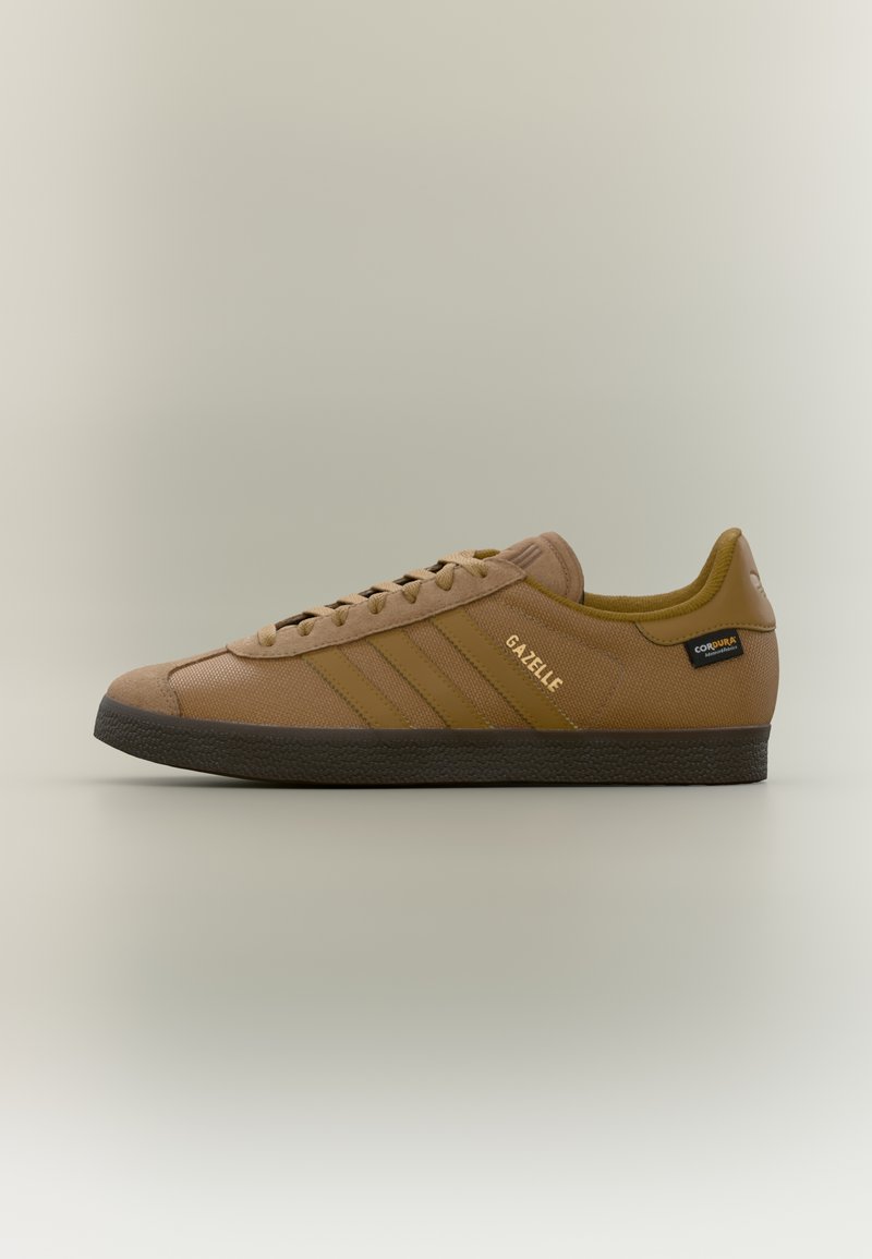 Καφέ αθλητικό παπούτσι Adidas Gazelle με υφασμένο πάτο, τρεις ρίγες στο πλάι και ετικέτα από ύφασμα Cordura στη φτέρνα, σε απλό φόντο.