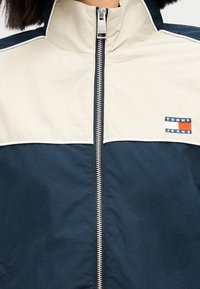 Μπλε σκούρο και κρεμ μπουφάν με φερμουάρ και ψηλό γιακά, με λευκή ρίγα και μικρό κεντητό λογότυπο "Tommy Jeans" στο στήθος.