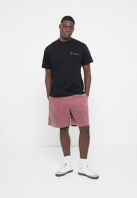 Svart bomull T-skjorte med logo foran, parret med rosa kordfløyelsshorts. Hvite joggesko med blå detaljer og hvite sokker. Enkel design.