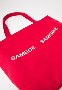Red cotton tote bag features two printed white logos "SAMSØE" on the front, with sturdy red fabric handles. Simple rectangular shape.