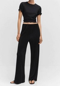 Femme portant un t-shirt noir court avec manches retroussées, un pantalon noir large taille haute et des sandales noires à bouts ouverts, debout devant un fond uni.