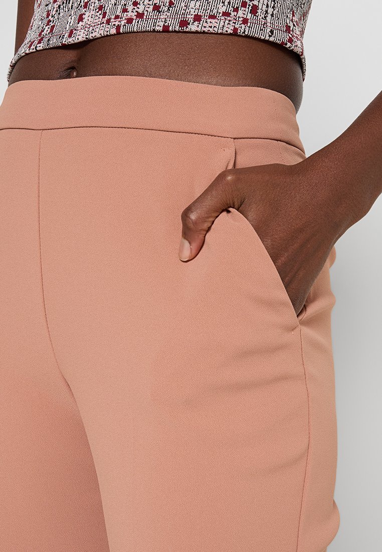 Jasnoróżowe, dopasowane spodnie z gładkiego materiału i bocznymi kieszeniami. Ręka modelki jest włożona do kieszeni, co podkreśla krój i fason.