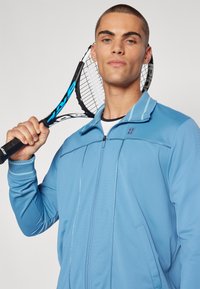 Chaqueta deportiva de color azul claro con cremallera y cuello alto, fabricada con un tejido texturizado y detalles a rayas. El modelo sostiene una raqueta de tenis.