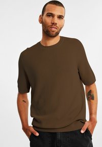Brun kortærmet strikket skjorte med et tekstureret mønster og rund hals. Modellen har sorte bukser på og viser en farverig tatovering på underarmen.