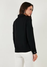Black long-sleeve blouse with a collar, featuring a smooth texture, button accents on sleeves, and a slightly curved hemline.