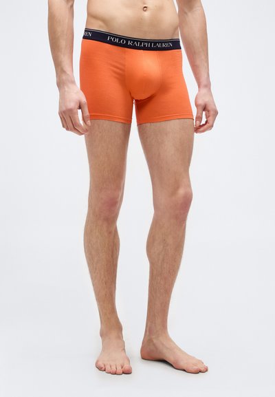Άνδρας μοντέλο φοράει πορτοκαλί ανδρικά εσώρουχα boxer με μαύρη ζώνη Polo Ralph Lauren, στέκεται ξυπόλυτος σε λευκό φόντο.