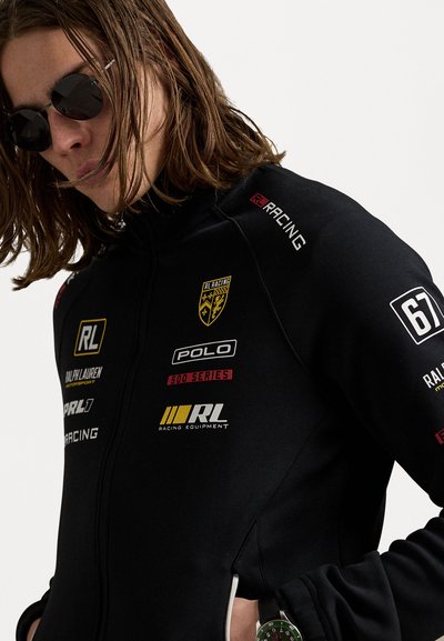 Schwarze Rennjacke mit Logos in Weiß, Gelb und Rot. Vollzipper, taillierte Passform, mit Reißverschlusstaschen und einer glatten Textur. Dünner Kragen.