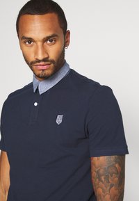 Navyblå polo-shirt med denimkrage, prydd med en liten broderad logotyp på vänster bröst. Mjukt, strukturerat tyg med korta ärmar.