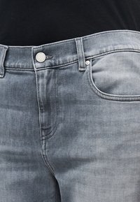 Gros plan de la taille d'un jean denim gris clair, montrant le bouton, les passants de ceinture et la poche avant avec un léger effet délavé et des détails de couture.
