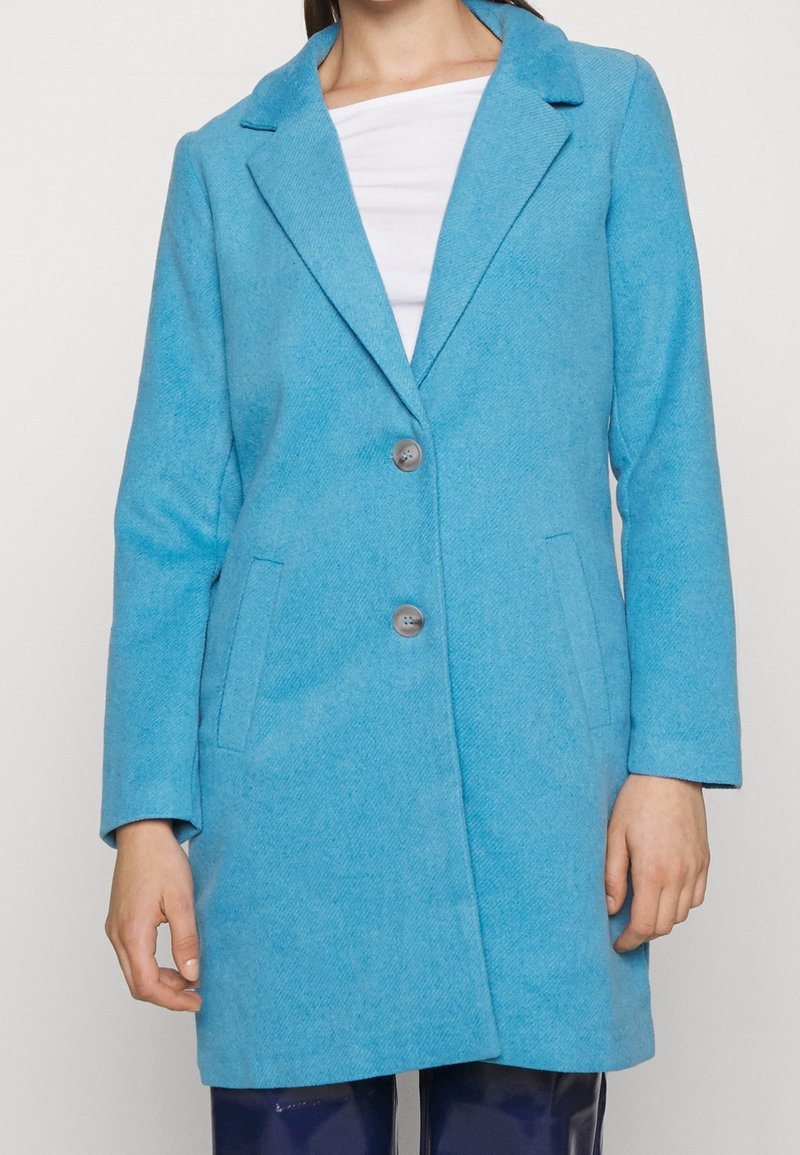Femme portant un manteau en laine texturé bleu vif avec deux boutons et des poches passepoilées par-dessus un haut blanc et un pantalon foncé.