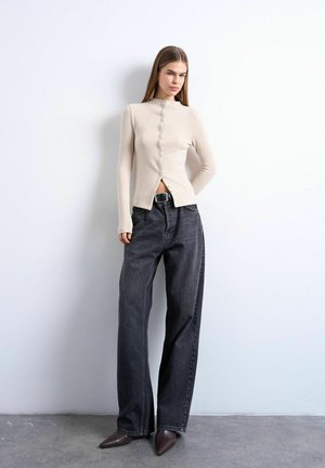 Kvinde står op ad en hvid væg iført beige knappet cardigan, mørke jeans med vide ben og sort bælte samt brune spidse sko.