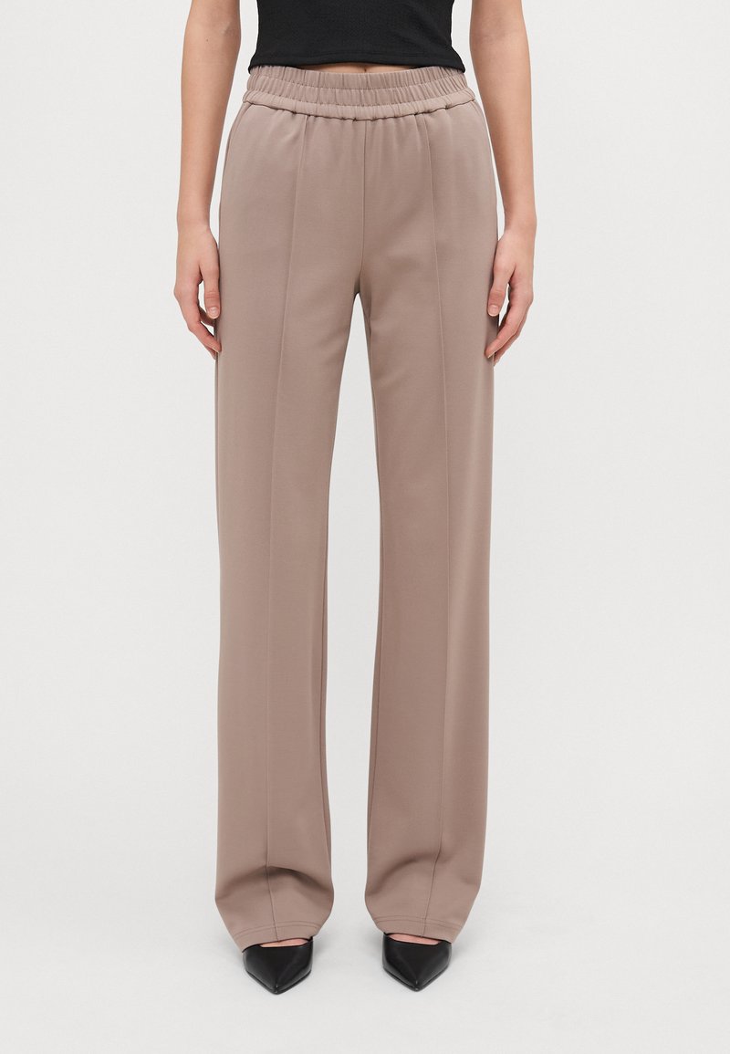 Beige wijde broek van zachte stof met een elastische tailleband en een plooi-detail. Gecombineerd met zwarte puntige hakken.