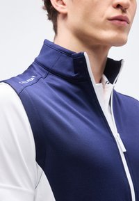 Κοντινό πλάνο ατόμου που φοράει γιλέκο με φερμουάρ σε σκούρο μπλε χρώμα πάνω από λευκό πουκάμισο, με ψηλό γιακά και το λογότυπο "Callaway" στον ώμο.