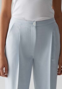 Pantalon bleu clair en tissu texturé, avec des plis, une poche latérale et une fermeture à bouton au niveau de la taille. Fini lisse.