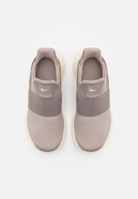 Zapatillas color marrón claro con banda elástica, parte superior de tela suave, cuello acolchado y suela de goma blanca; presenta un logo en el lateral.