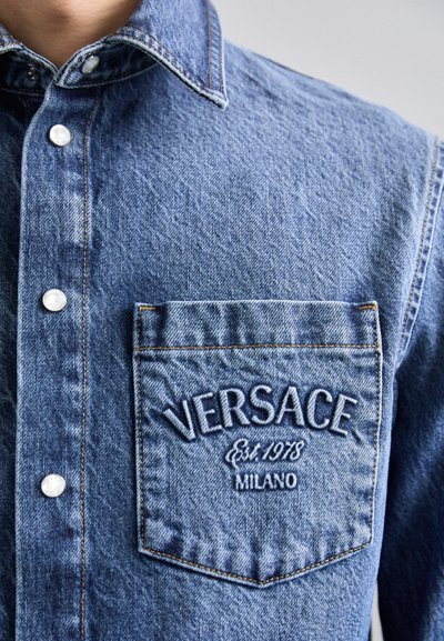 Camicia di denim blu chiaro con colletto classico, bottoni a pressione e taschino sul petto caratterizzato da ricamo "VERSACE" e "Est. 1978 MILANO."