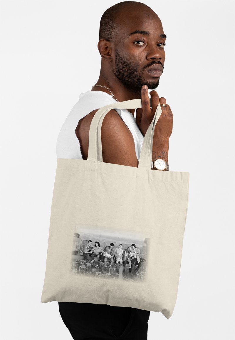 Uomo con barba e orologio che porta una borsa tote beige con una foto in bianco e nero di sei persone sedute su una trave d'acciaio sopra una città.