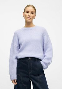 Jasnofioletowy sweter z miękkiego, puszystego materiału z okrągłym dekoltem i opuszczanymi ramionami, zestawiony z ciemnymi szerokimi spodniami dżinsowymi.