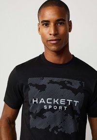 Hombre vistiendo una camiseta de cuello redondo negra con un patrón de camuflaje y el texto "HACKETT SPORT" en el frente, contra un fondo ligero y liso.