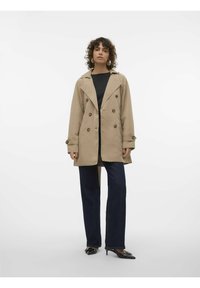 Beige trenchcoat med dubbelknäppt design, stora knappar och breda slag. Kombinerat med mörkblå raka jeans och svarta skor.