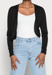 Schwarze, kürzere Strickjacke aus weichem Stoff mit langen Ärmeln und lockerer Passform, kombiniert mit hoch taillierten hellblauen Jeans.