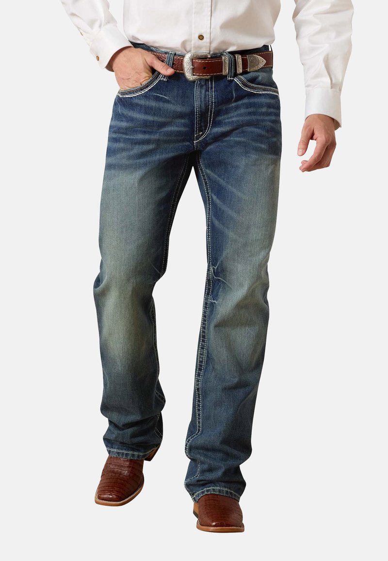 Man die versleten blauwe spijkerbroek draagt, een bruine leren riem met zilveren gesp, een wit shirt en bruine cowboylaarzen, staand met één hand in zijn zak.