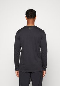 Mann mit kurzen, lockigen Haaren, der ein schwarzes langärmliges Shirt und schwarze Hosen trägt, stehend mit dem Rücken zu einem schlichten weißen Hintergrund.