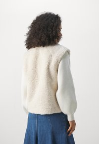 Kamizelka z sztucznego futra w kolorze kremowym, z textured powierzchnią, pod którą znajduje się biały, prążkowany rękaw. Niebieska spódnica dżinsowa dopełnia całość.