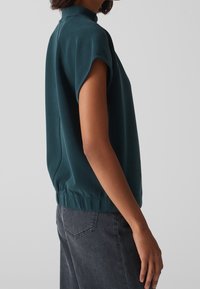 Maglietta a maniche corte color verde petrolio con collo alto, vita elastica e tessuto liscio, abbinata a jeans grigio scuro con cuciture minimaliste.