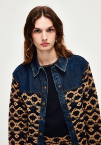 Jeansjacka med mörkblå överdel och en brun mönstrad underdel med geometriska mönster, avslutad med guldfärgade knappar.