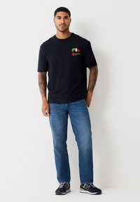 Melna kokvilnas T-krekls ar krāsainu Grinča attēlu uz krūtīm, kopā ar zilām džinsa biksēm un melnām sporta kurpēm.