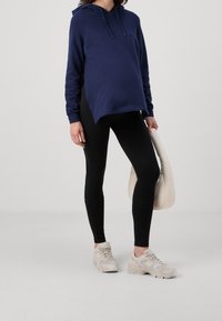 Marinblå huvtröja med rundad nederkant, dragsko på huvan och långa ärmar, kombinerad med svarta leggings och ljusbeige sneakers, håller en krämfärgad väska.