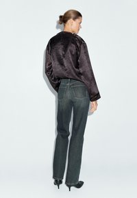 Donna con capelli raccolti in uno chignon, che indossa un top lungo e lucido di colore scuro, jeans grigi a gamba dritta e stivali neri con tacco, in piedi davanti a uno sfondo bianco.