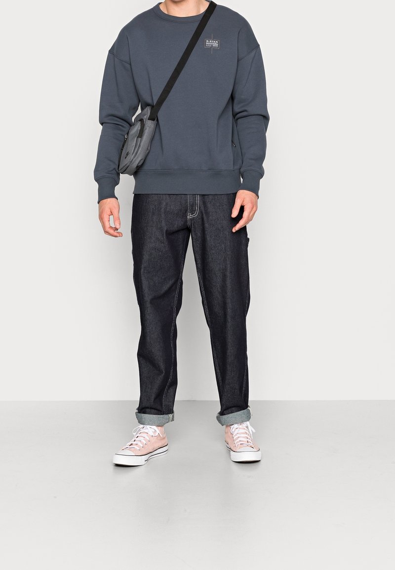 Mörkgrå sweatshirt med ribbad mudd, kombinerad med upprullade mörka jeans och ljusrosa sneakers. Liten logotyp på bröstet.