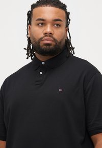 Čierne polo tričko s jemnou textúrou, zapínaním na dva gombíky a malým vyšívaným logom na ľavej strane hrude.