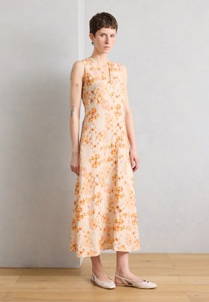 Noor naine lühikeste juustega, kandes varrukateta, virsikuvärvilist lillelist maksikleiti ja heledaid beežika slingback-tossusid, seisab ühe sirge seina ääres.