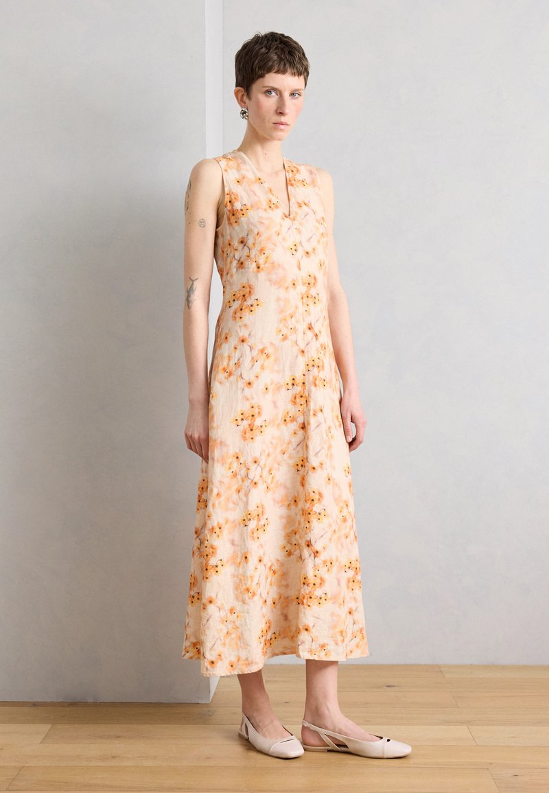 Νεαρή γυναίκα με κοντά μαλλιά, που φορά ένα αμάνικο μακρύ φόρεμα σε ροδακινί χρώμα με floral σχέδιο και ανοιχτά μπεζ παπούτσια με λουράκι, στέκεται μπροστά σε έναν απλό τοίχο.