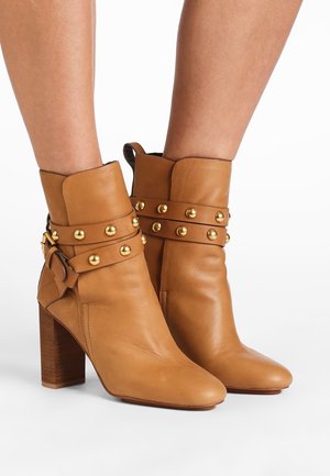 Bottes basses en cuir fauve avec talons blocs, clous décoratifs dorés sur deux sangles, portées sur des jambes nues sur un fond blanc.